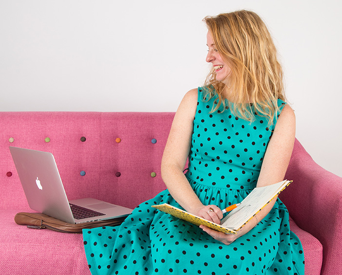Owner of Black Bee Creative, Steph Cronin-Milne wears a teal dress with black dots, sitting on a pink sofa looking at her laptop and holding a notepad.