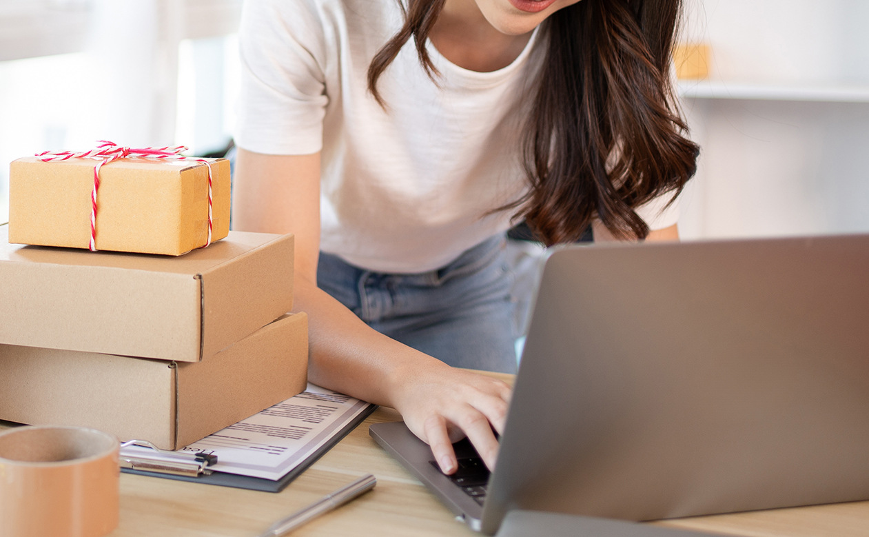 A woman typing on a laptop, with boxes, packing tape and a clipboard on the desk next to her.